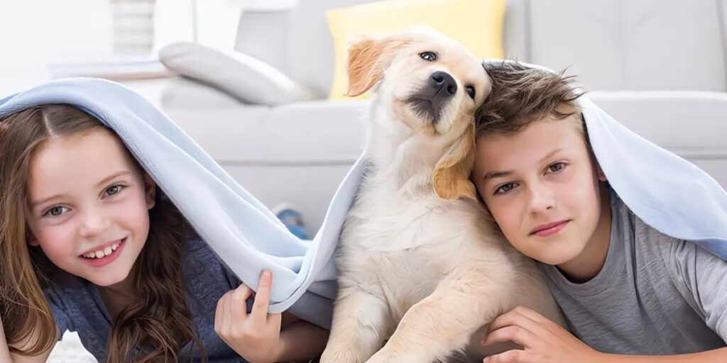 Two children and a playful dog peeking out from under a cozy blanket inside a home, symbolizing a warm, pet-friendly family environment.